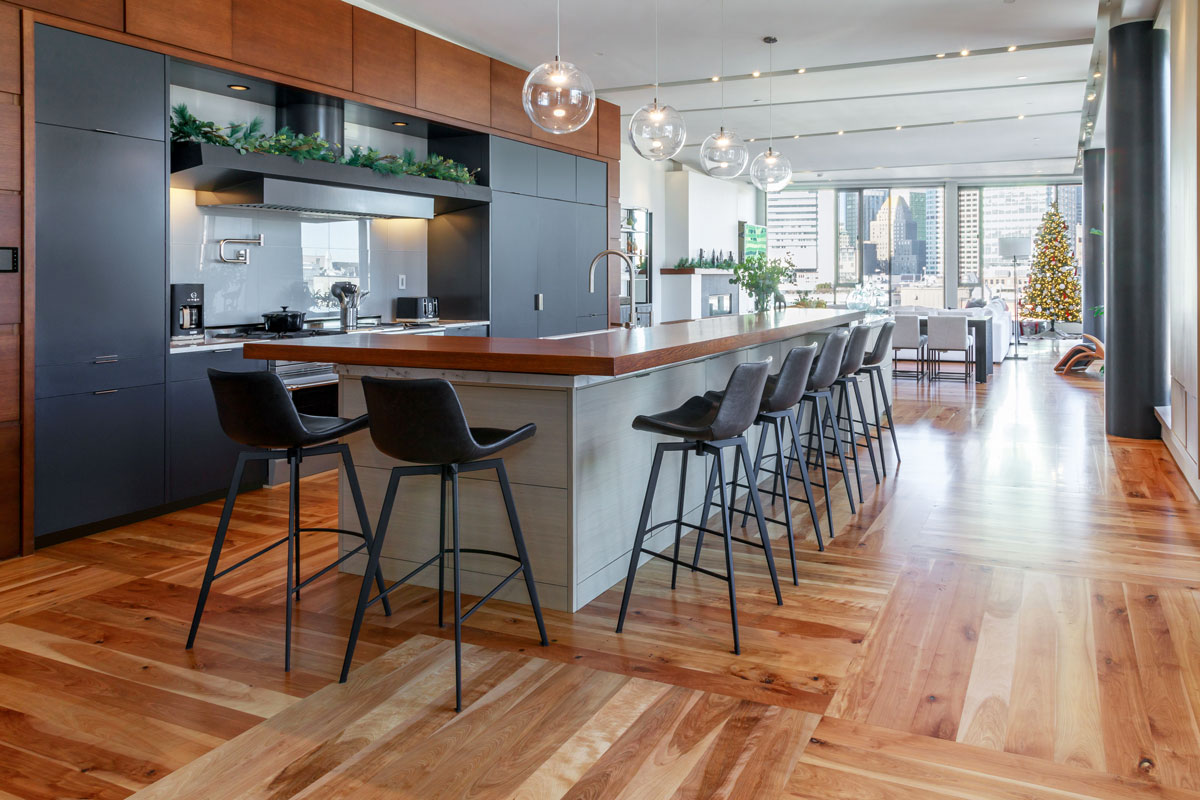 Modern kitchen with a wooden island, black stools, and dark cabinets, viewed against a backdrop of cityscape visible through large windows.